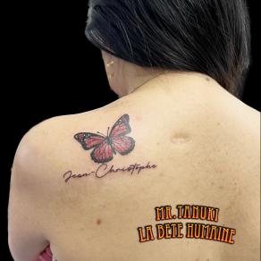 Tatouage de papillon sur l’omoplate assorti du prénom calligraphié Jean-Christophe, réalisé par Peter Galt